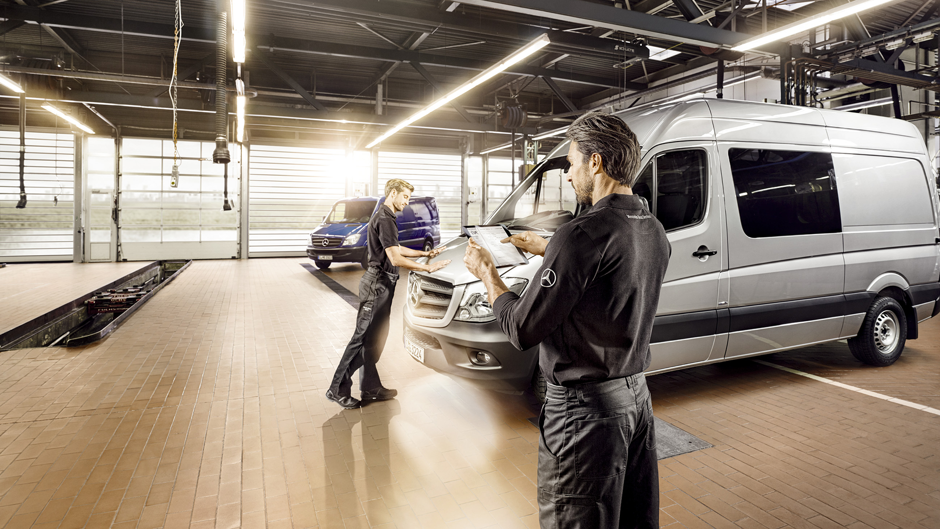 Kaksi Mercedes-Benz-mekaanikkoa huoltohallissa, jossa hopeanharmaa sekä sininen Mercedes Sprinter.