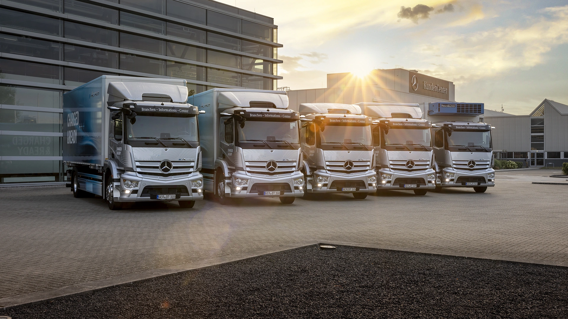 Priešais pastatą penki visiškai elektriniai Mercedes-Benz sunkvežimiai.